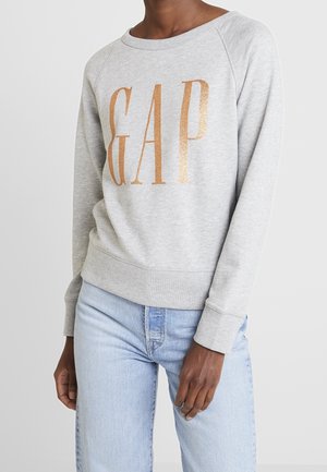 Person wearing light gray sweatshirt with large metallic gold "GAP" text and light blue denim jeans, standing against a plain background.