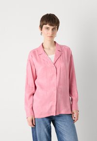 Pink, long-sleeve button-up shirt featuring a subtle texture and a relaxed fit, paired with blue jeans.