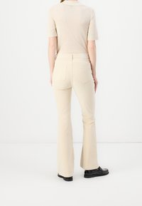 Pantalon beige évasé en coton doux, doté de deux poches arrière et d'une taille mi-haute. Associé à un haut beige clair et des chaussures noires.