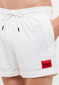 Balti maudymosi šortai su elastinga juosmens juosta, sutraukiamuoju virvute ir juodais antgaliais. Raudona etiketė su "HUGO" logotipu kairėje pusėje. Sklandus, lengvas audinys.