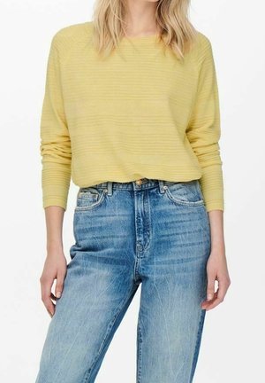 Un pull long à manches longues, texturé, de couleur jaune clair, associé à un jean bleu délavé taille haute. La tenue a une coupe décontractée et un style informel.