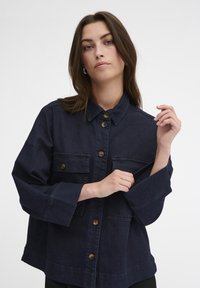 Giubbotto di jeans blu scuro, dotato di colletto, chiusura con bottoni, due tasche sul petto e maniche larghe con polsini cuciti.
