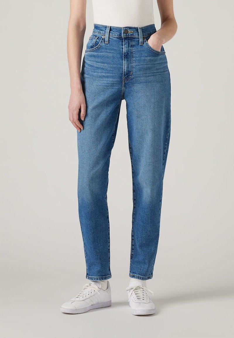 Personne portant un jean bleu taille haute avec des poches avant, associé à des chaussettes blanches et des baskets blanches à lacets sur un fond uni.