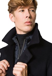 Czarny wełniany płaszcz z wysokim kołnierzem, nakładany na czarną zapinaną na zamek wewnętrzną kurtkę. Gładka tekstura, dopasowany krój i elegancka sylwetka.