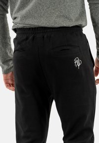 Pantaloni da jogger neri con vita elastica, tasche posteriori visibili e dettaglio del logo bianco. Tessuto morbido e strutturato con una vestibilità comoda.