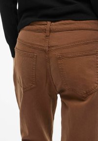 Bruine denimbroek met een rechte pasvorm, voorzien van twee achterzakken en zichtbare stikseldetails. Het materiaal lijkt zacht en textuur te hebben.