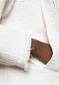 Biała, teksturowana tkanina z postrzępionymi brzegami; zbliżenie na rękaw i kieszeń, prezentujące tkany wzór i lekko uniesiony design.