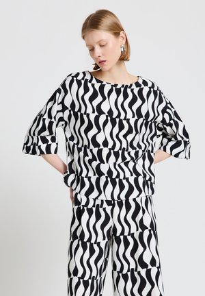 Femme aux cheveux courts portant un haut ample à motif ondulé noir et blanc et un pantalon assorti, mains sur les hanches, regard vers le bas.