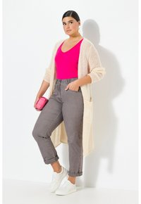 Langer, offen gestrickter Cardigan in Creme über einem figurbetonten pinken Top. Graue, hoch taillierte Jeans mit umgekrempelten Säumen und weißen Sneakers. Kleine pinke Handtasche.