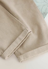 Manche en tissu beige avec coutures visibles et détail de poignet plié sur une surface claire avec un objet rayé partiellement visible en arrière-plan.