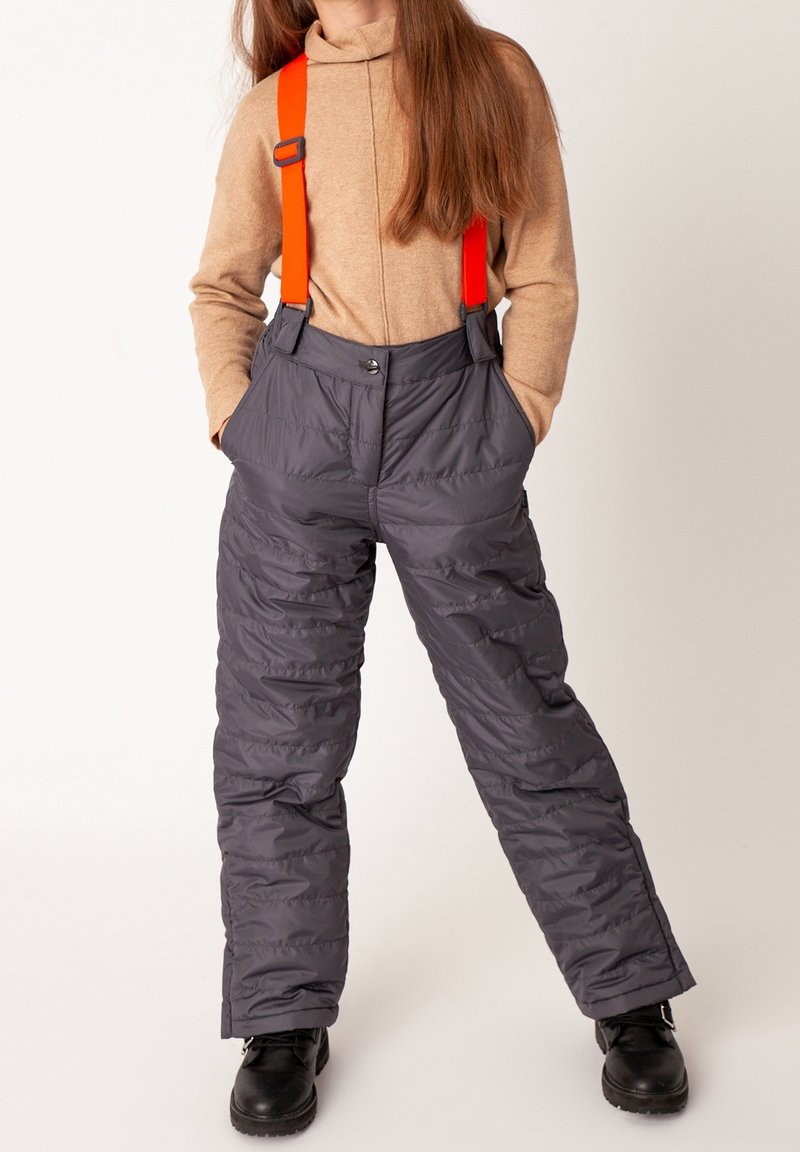 Barn iført lysebrun rullekravesweater og grå quiltede snekbukser med klare orange seler og sorte støvler, hænderne i lommerne.