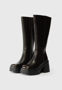 Μαύρες δερμάτινες μπότες μέχρι το γόνατο με χοντρή πλατφόρμα και στρογγυλή μύτη, διαθέτοντας μια διακοσμητική φερμουάρ στο πίσω μέρος.