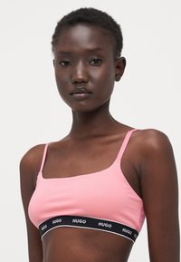 Bralette en tissu rose avec des bretelles réglables et une large bande élastique noire arborant "HUGO" en lettres blanches sur le bas.
