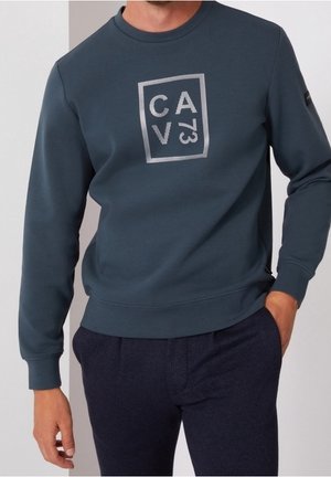 Mand iført en mørkeblå langærmet sweatshirt med et hvidt rektangulært logo med bogstaver og et nummer på brystet.