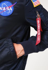 Sinise bomber-jakk, mis on valmistatud siledast nailonist, sisaldab NASA paanot, ribakleepidega varrukaid ning punast "EEMALDA ENNE LENDU" sildikest varrukal.