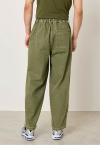 Olivově zelené kalhoty s širokými nohavicemi a elastickým pasem, se jednou zadní kapsou a měkkou, strukturovanou látkou. Nosit s stříbrnými teniskami.