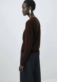 Pull en tricot marron à manches longues, avec des poignets et un ourlet côtelés ; associé à un pantalon large gris foncé. Boucles d'oreilles géométriques en argent en accent.