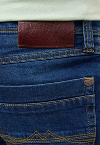 Granatowe dżinsy charakteryzują się brązową skórzaną łatą z napisem "MUSTANG", żółtym przeszyciem oraz unikalnym wzorem przeszyć na tylnych kieszeniach.