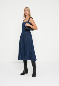 Donkerblauwe denim A-lijn jurk met verstelbare bandjes, zichtbare stiksels en een jurk tot op de knie; gecombineerd met zwart kniehoge laarzen en een zwarte tas.
