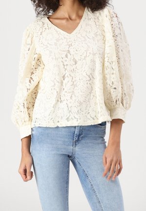 Kobieta ubrana w kremową, koronkową bluzkę w kwiatowy wzór z dekoltem w serek i bufkami na długich rękawach, w połączeniu z jasnoniebieskimi dopasowanymi dżinsami.