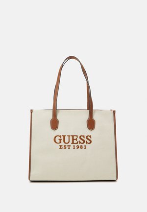 Beżowa torba typu tote z brązowymi skórzanymi uchwytami i wykończeniami, z haftem "GUESS EST 1981" w kolorze brązowym na środku przodu.
