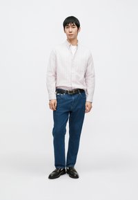 Lyserosa stripete skjorte med knapper, mørkeblå denimjeans, svart belte og svarte sko. Modellen står foran en ensfarget hvit bakgrunn.