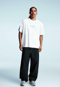 Tricou alb din bumbac cu imprimeu albastru "Value", asortat cu pantaloni largi din denim negri și adidași gri cu accente albe.