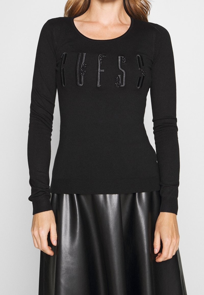 Femme portant un pull noir ajusté avec le lettrage "GUESS" orné et une jupe en simili cuir noir, debout devant un fond uni.