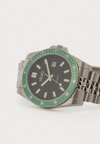 Kvartsur med svart urtavla, grön bezel, armband i rostfritt stål och silverfärgade timmarkörer. Har ett datumfönster vid klockan tre.