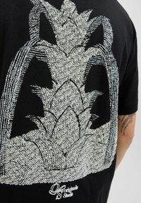 Camiseta negra con un gráfico blanco de un diseño de piña en la parte posterior. La textura parece tejida, con una etiqueta de marca en la parte inferior.