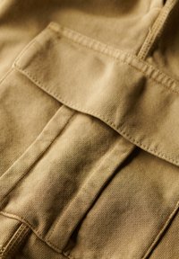Tejido beige con un acabado texturizado, que presenta un gran bolsillo de parche y detalles de costura visibles, adecuado para uso casual o de trabajo.