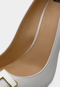 Weißer High Heel aus Leder mit geschlossenem Zehenbereich, ausgestattet mit einer rechteckigen Schnalle in Gold und Weiß sowie einer glatten, braunen Innensohle.