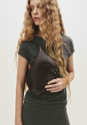 Geantă din piele maro, cu formă rotundă, prezentând capse argintii în colțuri. Modelul poartă un top și o fustă gri pe corp.