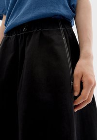 Zwarte broek met wijde pijpen, een elastische tailleband en een trekkoord. Heeft witte stiksels en zijzakken. Zachte katoenen stoftextuur.