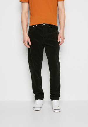 Mand iført sorte kordbuksere, orange kortærmet skjorte og hvide sneakers, der står mod en ensfarvet hvid baggrund.
