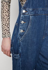 Denim kombinezoni z oprijetim krojem, temno modre barve, srebrnimi gumbi in stranskimi žepi. Notranji sloj vključuje bež majico z črnimi leopardjimi lisami.