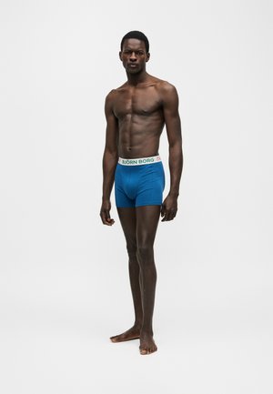 Hemdloser Mann steht barfuß auf weißem Hintergrund und trägt blaue Boxershorts mit weißem Björn Borg Bund.