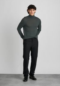 Tamsiai žalias golfas su lygia tekstūra, derinamas su juodomis kelnėmis ir juodais batais, pateiktas neutraliame fone.