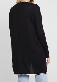 Černý pletený dlouhý kardigan s volnějším střihem, s bílým lemováním na spodním okraji a manžetách a svislým švem na zádech.