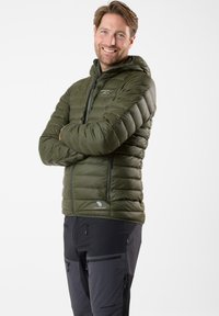 Oliven grøn, hættejakke med puf-effekt og lodret quiltet tekstur, lynlås lukning og premium dunlabel, parret med sorte tekniske bukser med lynlåslommer.
