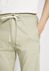 Helle grüne Hose mit einem Kordelzug an der Taille und silbernen Metallspitzen; verfügt über Seitentaschen und eine glatte Stofftextur.