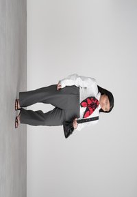 Γυναίκα με γκρι παντελόνι και άσπρο πουκάμισο, με μεγάλο κόκκινο και μαύρο κασκόλ, κρατά μαύρη τσάντα στον ανθισμένο γκρι φόντο.