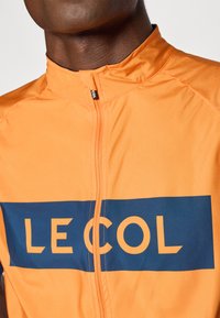 Oranžinė dviratininkų striukė su aukštu apykakle, su priekiu užtraukiama užtrauktuku ir jūrų mėlynos spalvos "LE COL" logotipu per krūtinę. Sklandžiai, lengvai medžiagai.