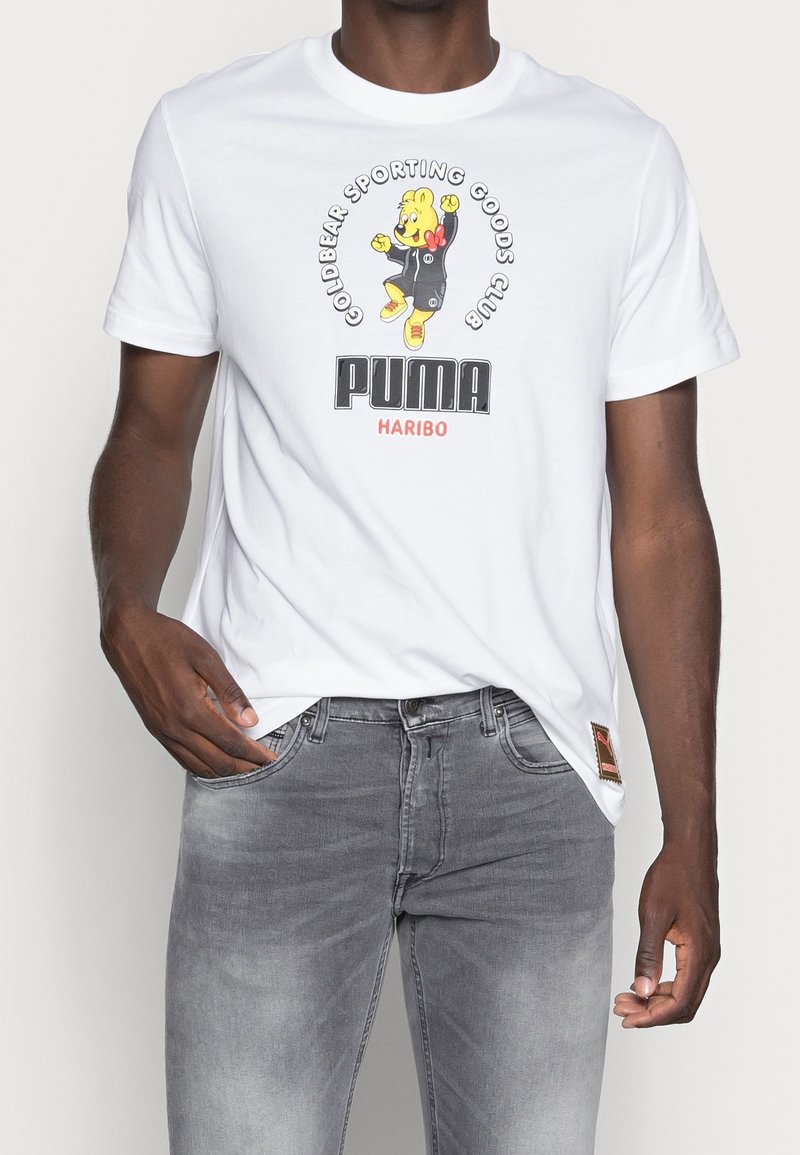 Camiseta blanca de algodón con un gráfico de un oso de dibujos animados que incluye el texto "PUMA" y "HARIBO". Combinada con unos jeans de mezclilla gris claro.