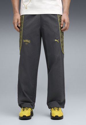 Person trägt schwarze, weit geschnittene Hosen mit gelben Pokémon- und Puma-Logos, gelben Seitenmustern sowie gelben und schwarzen Turnschuhen.