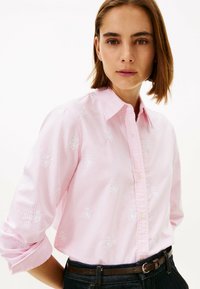 Light pink button-up shirt with white stripes and floral embroidery details. Features a classic collar and rolled sleeves. Worn with a dark belt.