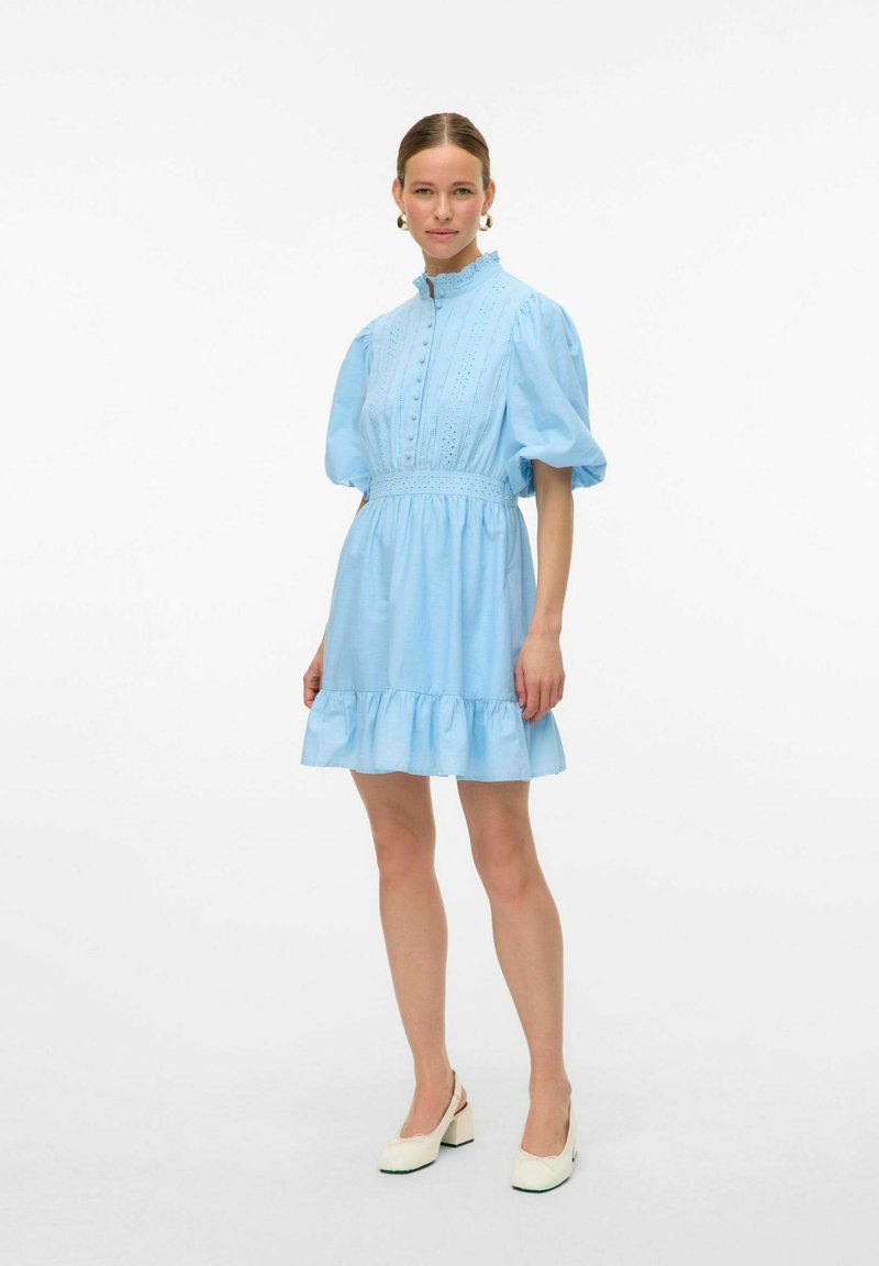 Svetlo modra kratka obleka z visokim ovratnikom, napihnjenimi rokavi, sprednjimi gumbi in nagubanimi robovi. Iz bombažne tkanine z vezenimi detajli.