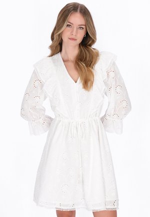 Młoda kobieta z długimi falującymi włosami, ubrana w białą sukienkę z ażurowego materiału, z bufkami, falbanami na ramionach i talią z troczkiem, stojąca z rękami na biodrach.