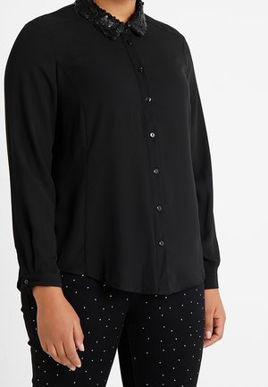 Chemise noire à col en sequins texturé, à manches longues et à coupe ajustée. Portée avec un pantalon noir clouté.