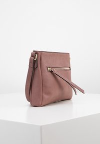 Pequeño bolso de piel malva cruzado con bolsillo con cremallera dorada y correa de hombro ajustable sobre una superficie blanca.